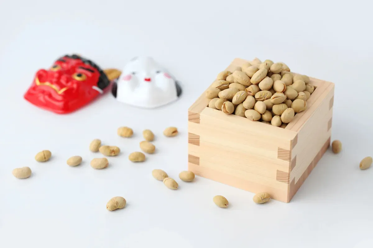 Wooden box filled with roasted soybeans used for Setsubun bean throwing, with oni masks in the background.