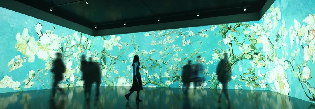 Visitors walking through a dark gallery surrounded by floor-to-ceiling digital projections of flowering branches in blue and white.
